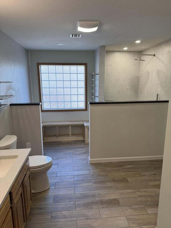 Remodeled bathroom with tiled walk in shower, wood look tile floor, vanity, toilet and glass block window