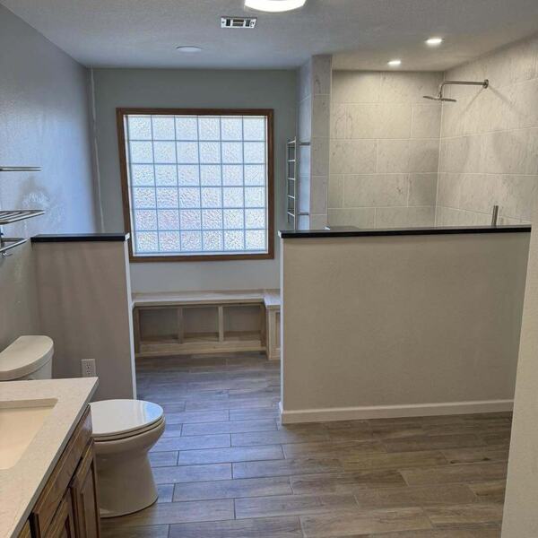 Remodeled bathroom with tiled walk in shower, wood look tile floor, vanity, toilet and glass block window
