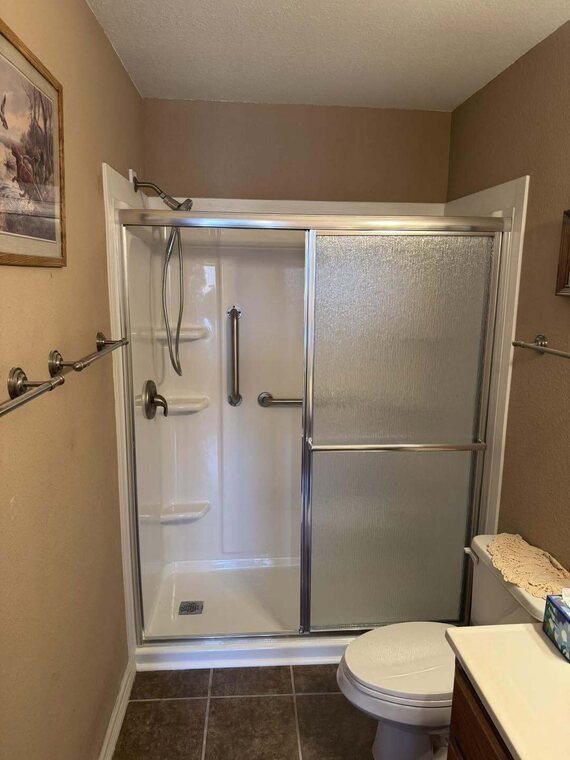 Walk in shower with sliding glass doors, grab bars, and a toilet in a tan walled bathroom with brown tile floor