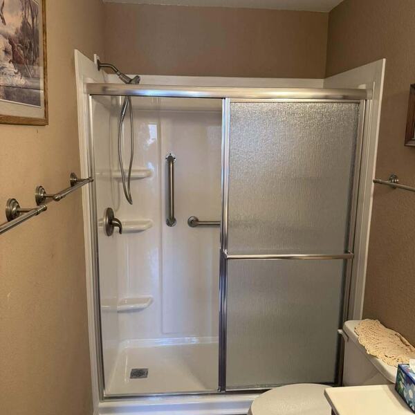 Walk in shower with sliding glass doors, grab bars, and a toilet in a tan walled bathroom with brown tile floor