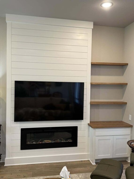 White shiplap accent wall with mounted TV, linear electric fireplace, and wood floating shelves over a cabinet
