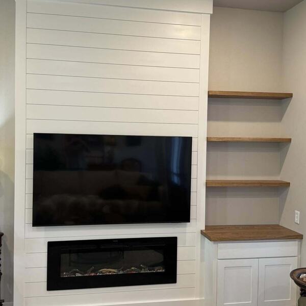 White shiplap accent wall with mounted TV, linear electric fireplace, and wood floating shelves over a cabinet