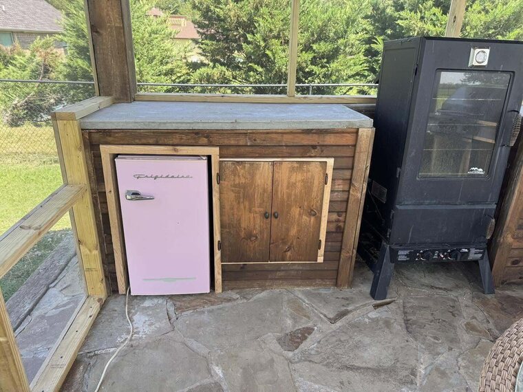 Wood cabinet built on a screened porch with a stone floor a small pink fridge and a black smoker