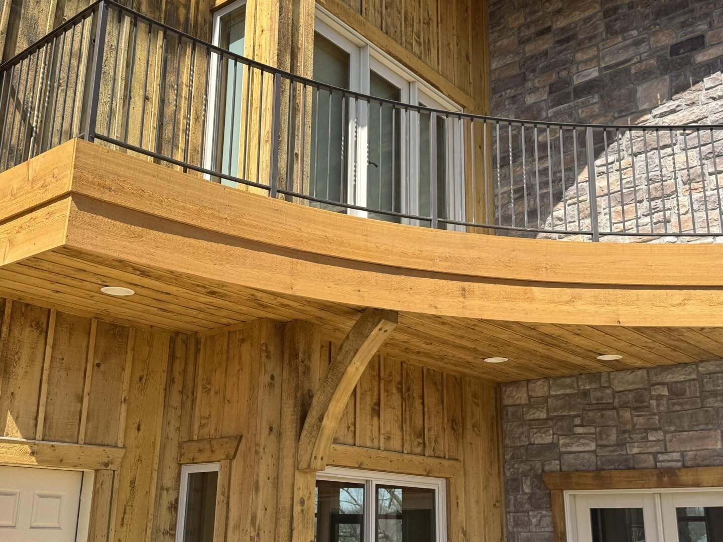 Curved wood balcony and covered porch ceiling with metal railing over stone and wood siding