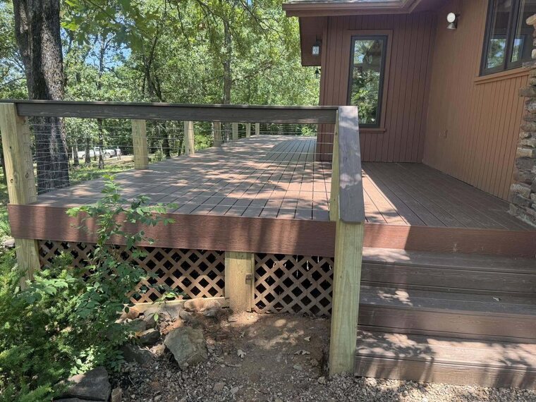 Wood deck with cable railing and lattice skirting beside a wood sided home in a wooded yard