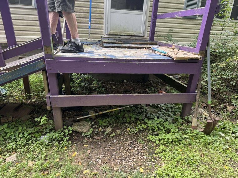 Worn purple painted wood deck with peeling boards and overgrown plants beside a house