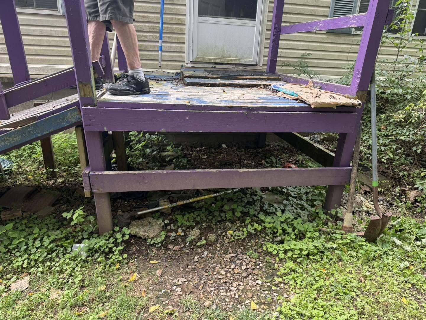 Worn purple painted wood deck with peeling boards and overgrown plants beside a house