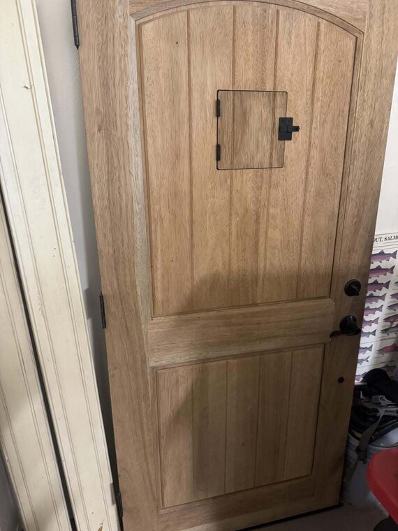 Unfinished wood plank door with an arched top, a small square access panel, and black hardware leaning in a garage