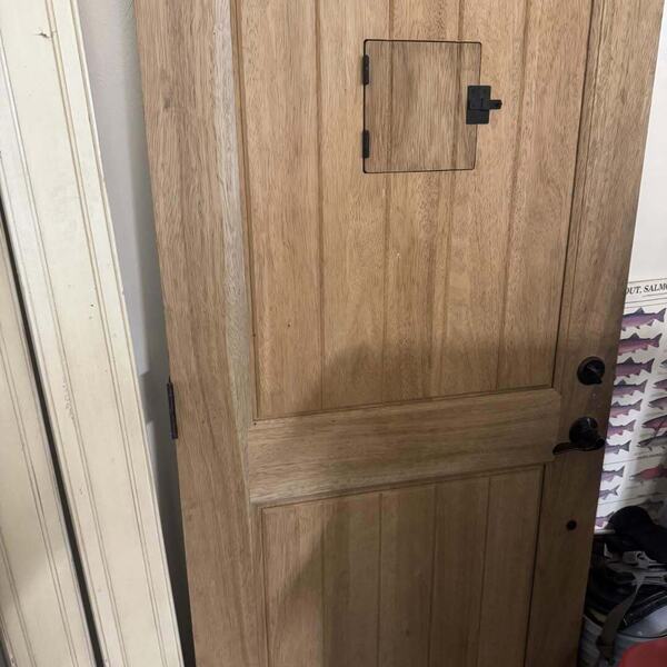 Unfinished wood plank door with an arched top, a small square access panel, and black hardware leaning in a garage