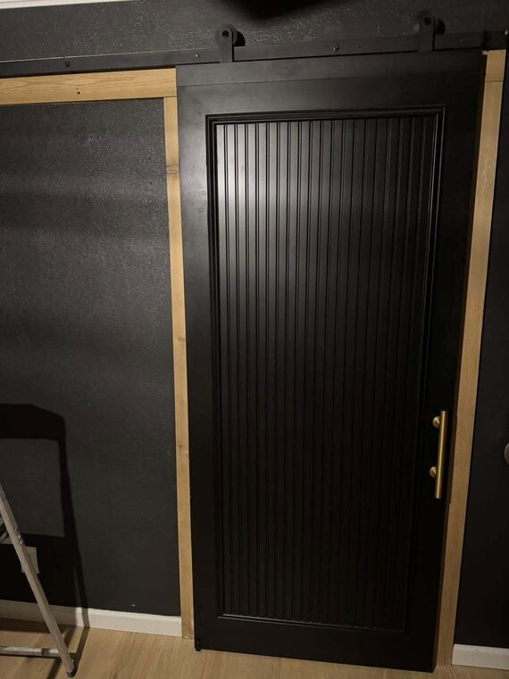 Black sliding barn door with vertical grooved panel and brass bar pull on black hardware over light wood floor