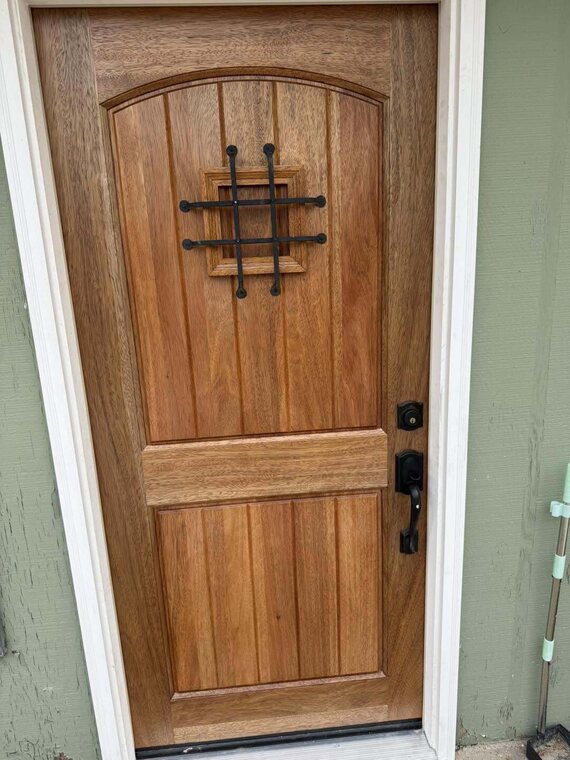 Stained wood exterior entry door with iron speakeasy grille and dark handleset on green wall