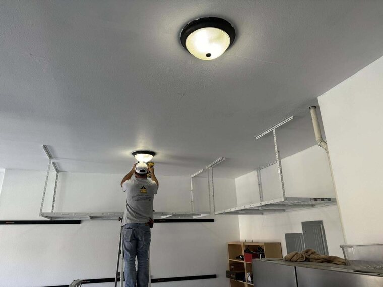Worker on a ladder installing a ceiling light in a garage with overhead storage racks