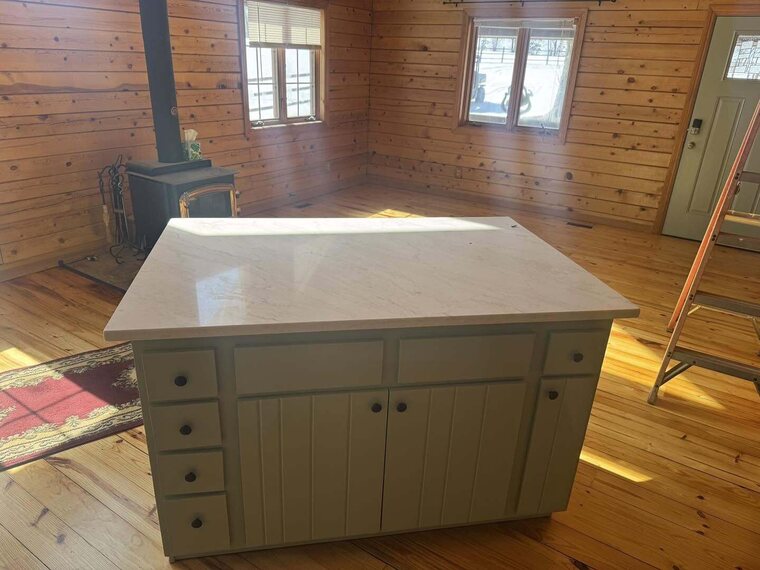 Kitchen island with sage green cabinets and a light stone top in a wood paneled room with a wood stove