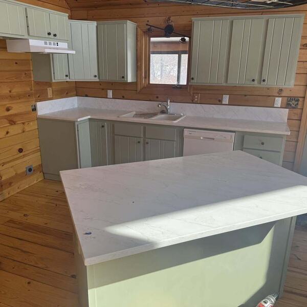 Cabin kitchen with sage green cabinets, light marble look countertops, sink, and white dishwasher in a wood paneled room