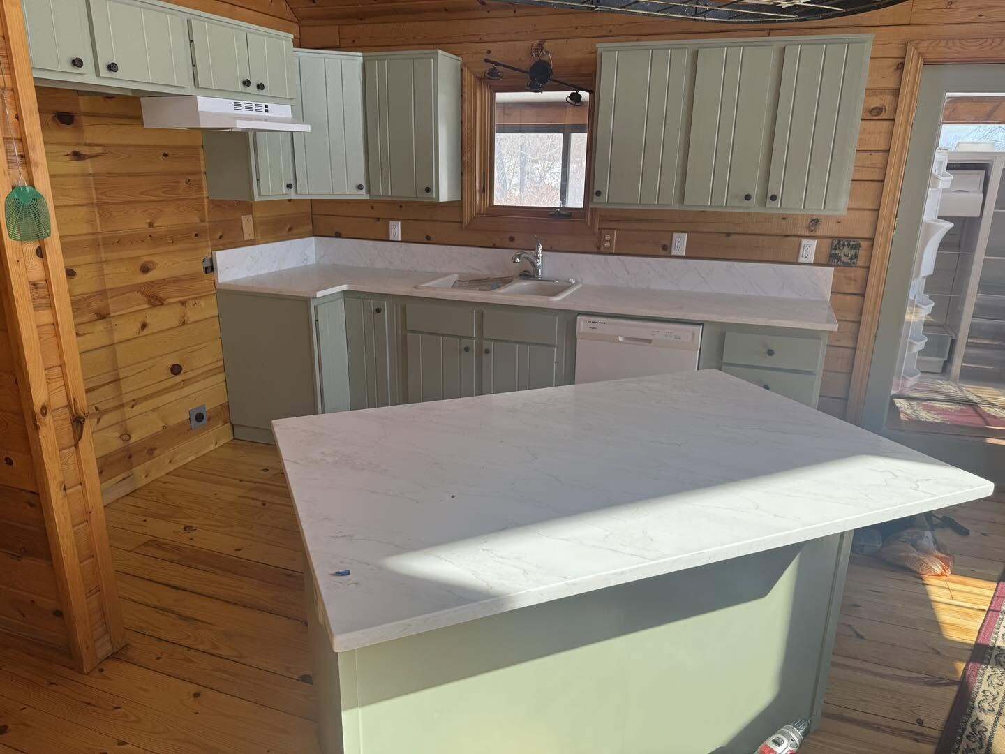 Cabin kitchen with sage green cabinets, light marble look countertops, sink, and white dishwasher in a wood paneled room