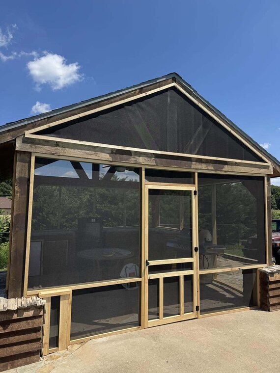 Wood framed screened porch with sliding screen door, stone base, and metal gable roof under blue sky
