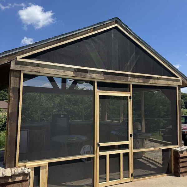 Wood framed screened porch with sliding screen door, stone base, and metal gable roof under blue sky