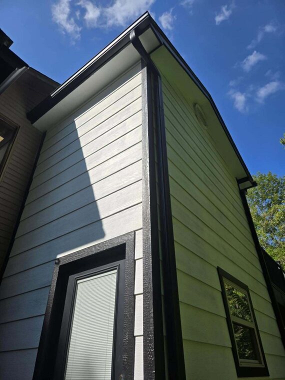 Exterior corner of a house with green and light gray lap siding, dark trim, gutter, and a window against blue sky