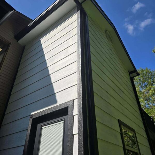 Exterior corner of a house with green and light gray lap siding, dark trim, gutter, and a window against blue sky