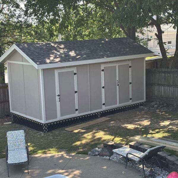 Gray storage shed with white trim, double doors, and a shingled gable roof in a fenced backyard