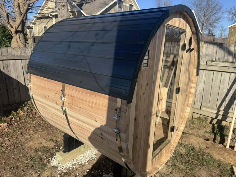 Cedar barrel sauna with a dark curved metal roof and glass door on a gravel base in a fenced backyard
