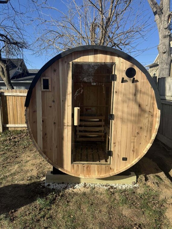 Cedar barrel sauna with a glass door on a gravel and wood base in a fenced backyard