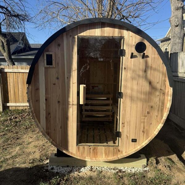 Cedar barrel sauna with a glass door on a gravel and wood base in a fenced backyard