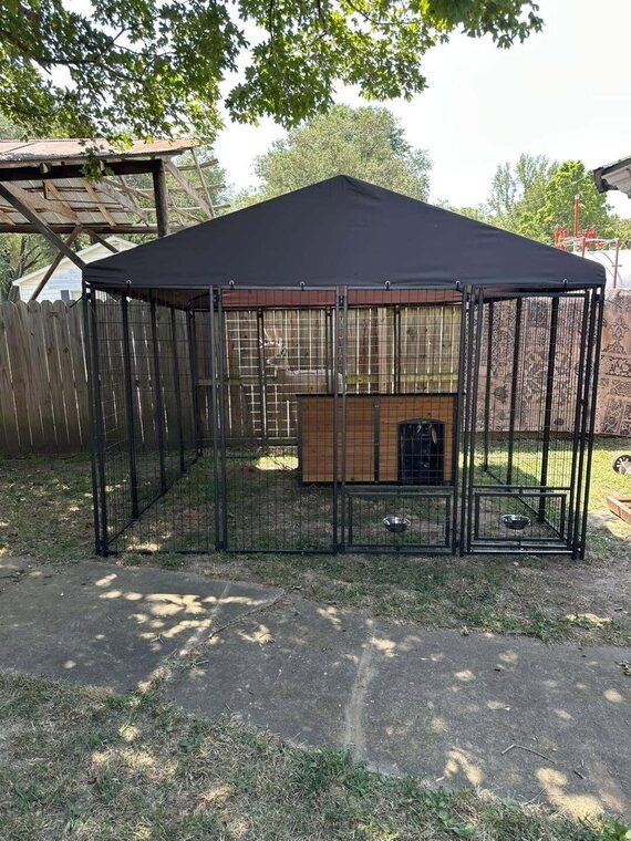 Outdoor metal dog kennel with a dark canopy roof and a wood dog house inside set on a yard