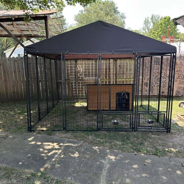 Outdoor metal dog kennel with a dark canopy roof and a wood dog house inside set on a yard