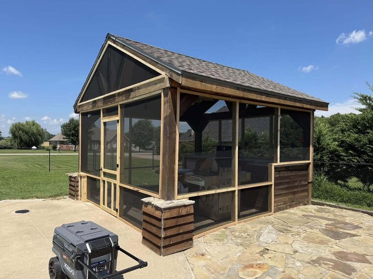 Wood framed screened structure with shingled gable roof on a flagstone patio with a lawn behind