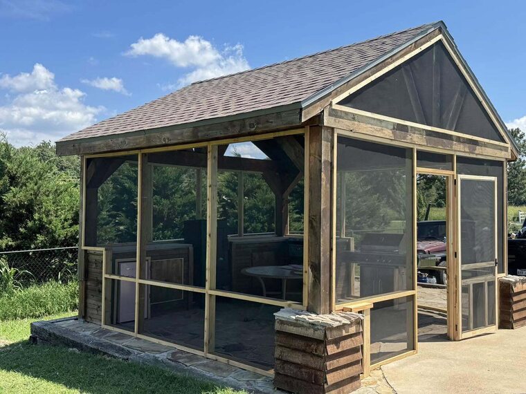 Wood framed screened structure with shingled roof and screen door beside an outdoor grill area