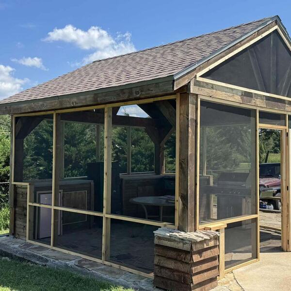 Wood framed screened structure with shingled roof and screen door beside an outdoor grill area